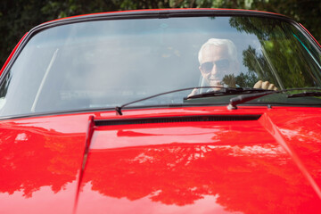 Senior man wearing aviator sunglasses driving red convertible featuring wipers on leafy driveway
