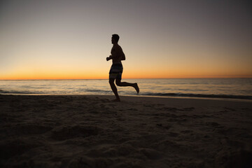 Fototapeta premium Shirtless man jogging along sandy shoreline by calm ocean at sunset wearing striped board shorts