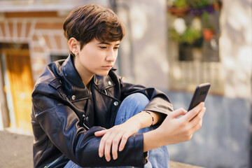 A young teenager with a smartphone is seated in an urban setting, surrounded by a lively cityscape