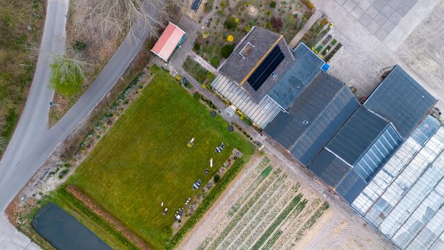 Aerial view of rural landscape with greenhouses, crop fields, canal, pasture with cows, wooded area, and residential neighborhood under a cloudy sky.