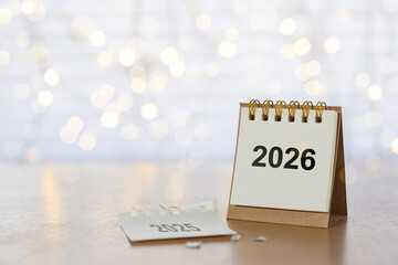 Desk calendar turning from 2025 to 2026 on a wooden table with warm bokeh lights, symbolizing year-end farewell, New Year celebration, resolutions, goals, and fresh beginnings with generous copy space
