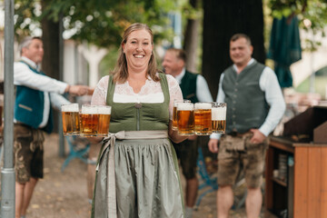 Cheerful waitress in Bavarian costume serving beer mugs to a group of friends in traditional attire enjoying Oktoberfest in a lively beer garden