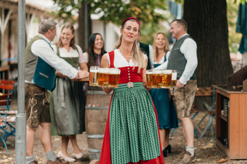 Happy Bavarian waitress in dirndl serving large mugs of beer to group of friends enjoying Oktoberfest atmosphere in traditional beer garden