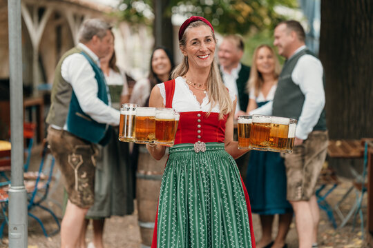 Cheerful waitress in Bavarian dirndl carrying beer mugs to frien