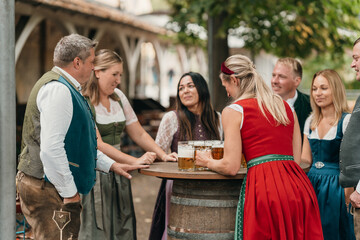 Smiling waitress carrying beer mugs to Bavarian dressed friends celebrating Oktoberfest in beer garden friendship leisure and festive culture