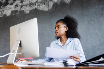 A young professional woman works at her desk with a computer, reviewing charts and opening a file folder, reflecting focus, organization, productivity, and dedication in a modern business environment.