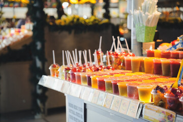 Colorful fruit juices, smoothies, and fresh fruit salads are displaying in plastic cups with straws and spoons at an indoor market stall, offering healthy and refreshing drinks