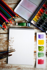 Handmade artistic workspace with blank white paper surrounded by brushes, pencils, paints and creative tools. Perfect background for art projects, mockups, invitations or design templates.