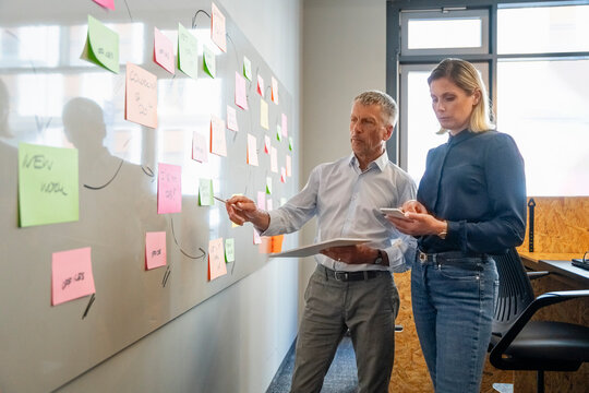 Senior businessman discussing over adhesive notes with colleague using smart phone in office