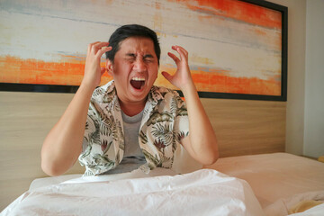 asian man painful and miserable sitting on bed angry and stress face with hands.