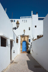 A narrow sun-drenched alley in Rabat Kasbah in Morocco, white walls and a blue arched door. Retro wall lanterns and small square windows under a clear blue sky. Quiet midday scene without people