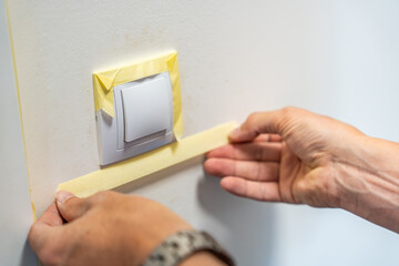Man covering a light switch with masking tape before painting the wall white. Home renovation, surface protection and careful preparation during interior work.