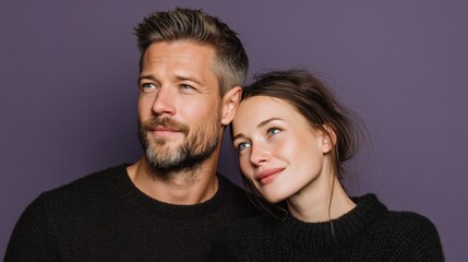 Fototapeta premium A young Caucasian man with a beard and a young Caucasian woman with brown hair pose closely together against a purple background. They appear happy and relaxed.