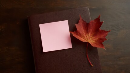Autumn Notes: A detailed overhead shot showcasing a closed journal, a vibrant red maple leaf, and a blank pink sticky note resting against each other on a dark wooden surface.