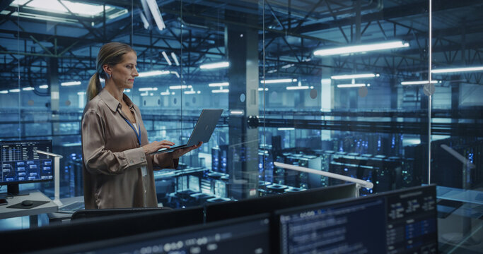 Caucasian Female Data Center Specialist Analyzes Server Performance on a Laptop, Optimizing Network Efficiency in a High-tech Environment. Developer Manages Cybersecurity and System Reliability