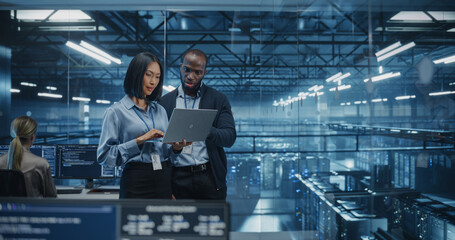 Male DevOps Engineer and Female IT Consultant Work on a Laptop Computer, Standing in an Office Above the Supercomputer with Server Cluster, Developing Automation Scripts for Artificial Intelligence