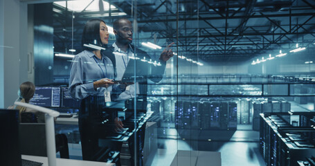 Two Diverse Data Center Engineers, a Black Male and a Asian Female, Collaborate Using a Laptop Computer in a Glass Office Next to a Futuristic Server Room, Analyzing Software Data on Screen