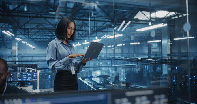 Asian Software Developer Analyze System Performance Metrics on a Laptop Inside a Modern Data Center. Young Female Manager Ensuring Efficient Operations for Critical Infrastructure - Powered by Adobe