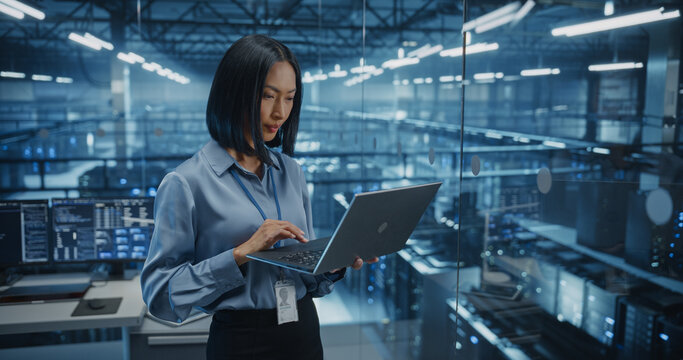 Positive Female Infrastructure Architect Reviewing Virtualization Strategies on a Laptop Computer, Standing in Office Next to Towering Racks of Servers, Planning to Scale Client Cloud Solutions
