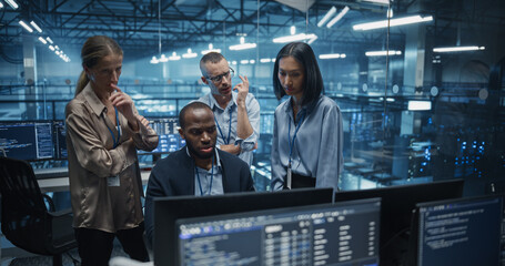 Diverse Team of IT Professionals Gathered Behind a Desk, Looking at Data on Computer Screens,...