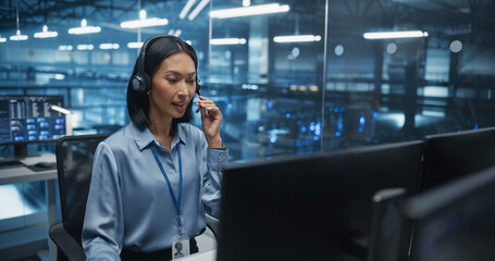 Wearing Headphones and Taking a Call, an Asian Female IT Specialist Focuses on Her Computer in the Data Center Office, Discussing System Updates and Performance Metrics with a Colleague