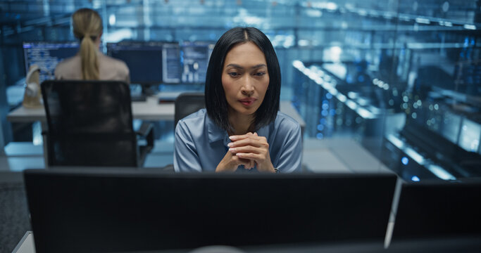 Beautiful Asian Female IT Engineer, Surrounded by Office Equipment, Studies Her Computer Display in a Modern Data Center, Reviewing System Health and Ensuring Optimal Server Operations - Powered by Adobe