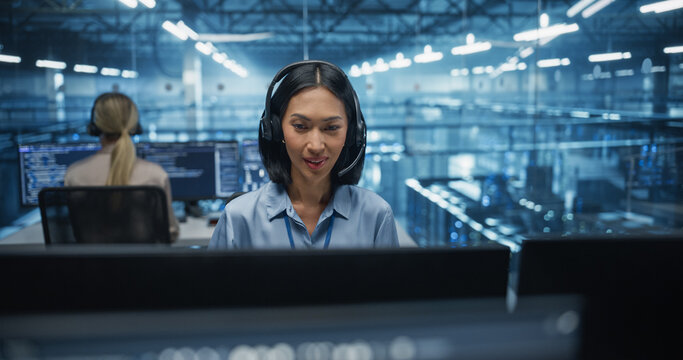 Wearing Headphones and Taking a Call, an Asian Female IT Specialist Focuses on Her Computer in the Data Center Office, Discussing System Updates and Performance Metrics with a Colleague