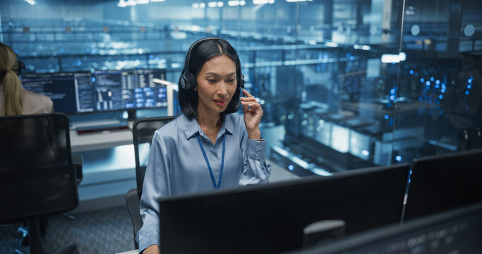 Focused and Professional, an Asian Female IT Analyst with Headphones Talks Through a Complex Network Configuration on Her Call, Reviewing Detailed Data on Her Desktop Computer in Data Center