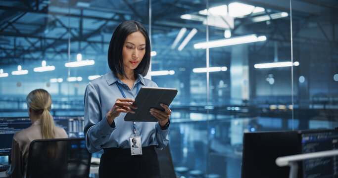 Beautiful Asian Female Cybersecurity Expert Works on Her Tablet Computer in a Modern Facility, Running Diagnostic Tests to Identify Network Vulnerabilities in a Data Center with Server Racks