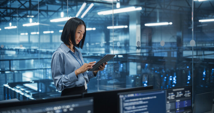 Asian Female Data Analyst Studies Online Logs on Her Tablet Computer, Standing in a Modern Data Center with Server Systems. Young Woman Searching for Patterns to Improve Online Data Flow