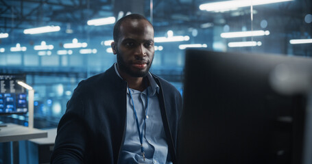 Handsome African Male IT Engineer, Surrounded by Office Equipment, Studies His Computer Display in a Modern Data Center, Reviewing System Health and Ensuring Optimal Server Operations
