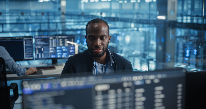 Handsome African Male IT Engineer, Surrounded by Office Equipment, Studies His Computer Display in a Modern Data Center, Reviewing System Health and Ensuring Optimal Server Operations