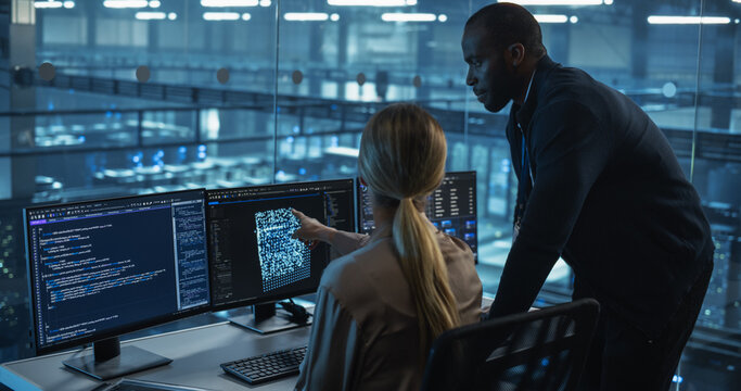 In a Data Center Office, a Female Software Developer Focuses on an AI Model Displayed Across Her Monitors, as a Black Colleague Stands Nearby, Offering Insightful Input for Their Project