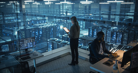 Female Cybersecurity Expert Standing with Her Back to Camera, Works on Her Tablet in a Modern Facility, Running Diagnostic Tests to Identify Network Vulnerabilities in a Data Center with Server Racks