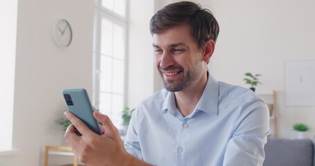 Excited young happy man using mobile phone sitting at home or in office. Male person looking at smartphone screen and making yes gesture reading pleasant sms message. 4k video. Slow motion video. - Powered by Adobe