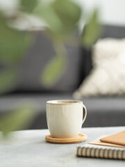 Ceramic coffee mug on wooden coaster placed on table with notebooks, in front of a sofa with cushions