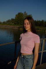 Fototapeta premium A young female model poses outdoors near a lake, enjoying the warm sunlight. She wears a fitted pink top and light blue jeans, with long brown hair flowing down. 