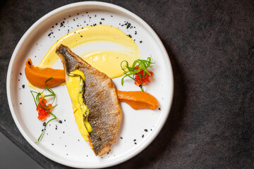 Gourmet plated fish dish with crispy skin, yellow sauce, orange puree swipes, fish roe, herbs, and black salt on white plate. Fine dining presentation on dark background.