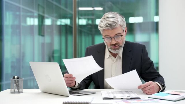 Confused puzzled mature businessman having difficulty with paperwork sitting at workplace in business office. Frustrated financier unhappy with bad financial results holding and reviewing documents
