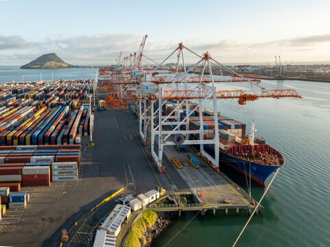 Tauranga, New Zealand - 24 February 2025: Aerial view of container ship berthed at port, where cranes tower over stacks of colorful containers near Mount Maunganui.