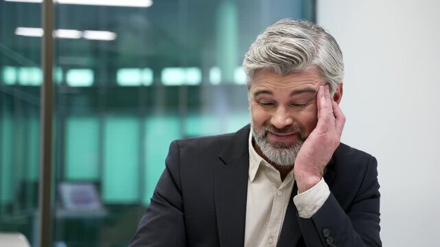 Mature bearded businessman suffering from headache sitting at workplace in business office. Unhealthy worker in formal suit holding and massaging his temples with his hands. He is exhausted. Close up