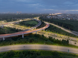 An aerial, wide-angle perspective of a highway interchange at dusk, with the curved ramps and overpasses illuminated, showing the smooth flow of traffic surrounded by trees and urban developments.