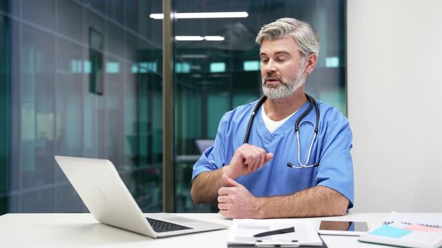 Confident mature doctor in blue coat talking on video call using laptop sitting in hospital office. Medical worker physician communicates remotely at conference or has online consultation with patient
