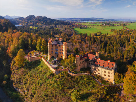 Aerial view of Hohenschwangau Castle perched atop a hill, surrounded by a tapestry of autumnal forests and distant lakes, Schwangau, Bayern, Germany.
