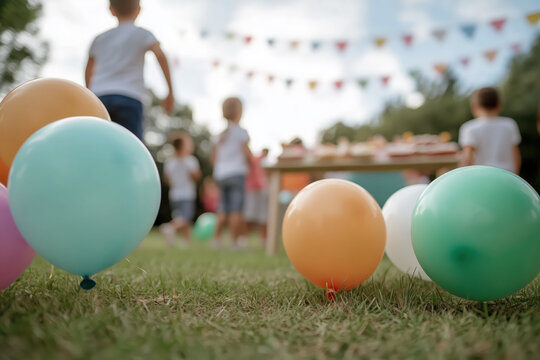 Children playing and celebrating a birthday party outdoors in a backyard, with colorful balloons lying on the grass and decorative flags hanging in the background