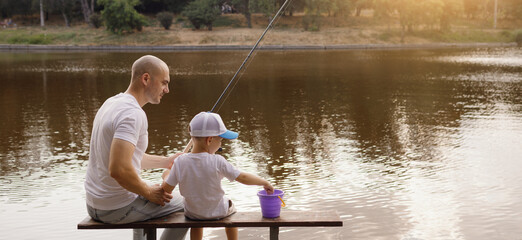 Father and young son fishing at calm lake in countryside at sunset. Horizontal family lifestyle photo with nature background and warm summer mood.