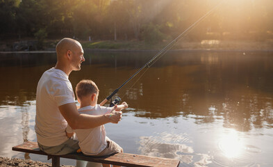 Father and young son fishing at calm lake in countryside at sunset. Horizontal family lifestyle photo with nature background and warm summer mood.