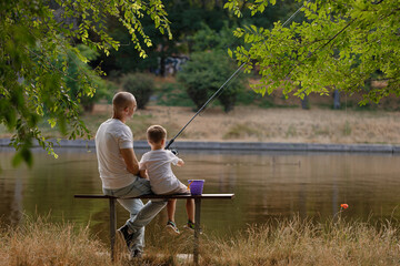 Father and young son fishing at calm lake in countryside at sunset. Horizontal family lifestyle photo with nature background and warm summer mood.