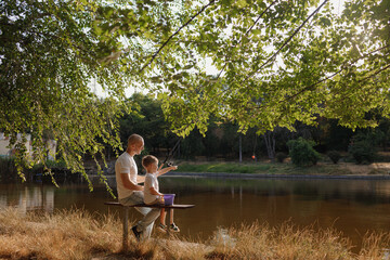 Father and young son fishing at calm lake in countryside at sunset. Horizontal family lifestyle photo with nature background and warm summer mood.