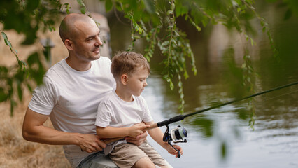 Father and young son fishing at calm lake in countryside at sunset. Horizontal family lifestyle photo with nature background and warm summer mood.